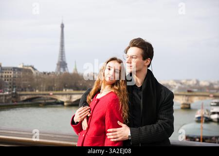 Ein lächelndes Paar umarmt sich auf einer malerischen Pariser Brücke mit Blick auf die seine mit dem Eiffelturm dahinter. Liebevolle Pose Fangen Sie einen zeitlosen romantischen Moment im Herzen Frankreichs ein. Stockfoto