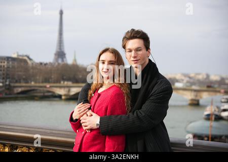 Ein stilvolles Paar umarmt sich auf einer Pariser Brücke und genießt einen romantischen Moment mit dem Eiffelturm und der seine im Hintergrund. Outfits ergänzen das malerische Stadtbild und symbolisieren die Liebe in Paris. Stockfoto