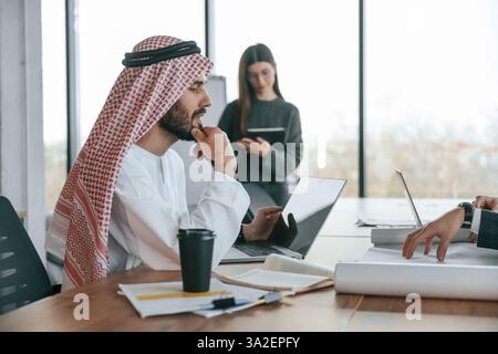 Seitenansicht. Muslimischer Geschäftsmann in traditioneller Kleidung mit Kollegen im Amt. Stockfoto