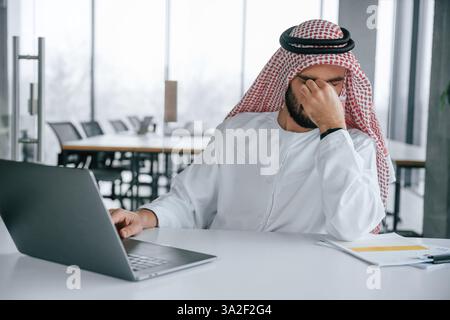 Müdigkeit. Mit Laptop am Tisch sitzen. Erfolgreicher muslimischer Geschäftsmann in traditioneller Kleidung in seinem Büro. Stockfoto