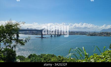 Ein atemberaubender Panoramablick auf eine große Brücke, die sich über ein glitzerndes Gewässer erstreckt und eine pulsierende Skyline der Stadt mit üppigen, grünen Hügeln verbindet. Stockfoto