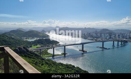 Ein atemberaubender Panoramablick auf eine große Brücke, die sich über ein glitzerndes Gewässer erstreckt und eine pulsierende Skyline der Stadt mit üppigen, grünen Hügeln verbindet. Stockfoto