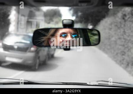 Die lächelnde Frau in Sonnenbrille blickt durch den Autospiegel zurück und blickt durch die Windschutzscheibe in Schwarz-weiß Stockfoto