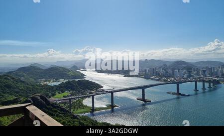 Ein atemberaubender Panoramablick auf eine große Brücke, die sich über ein glitzerndes Gewässer erstreckt und eine pulsierende Skyline der Stadt mit üppigen, grünen Hügeln verbindet. Stockfoto