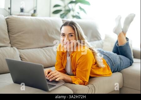 Die fröhliche hübsche weiße Frau in gelbem Hemd und Jeans liegt bequem auf einer Couch im Wohnzimmer, während sie online an ihrem Laptop arbeitet, einen produktiven Moment zu Hause genießt und in die Kamera lächelt Stockfoto