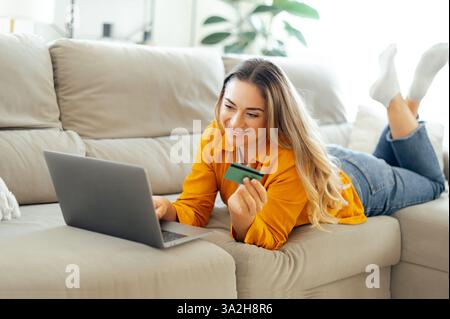 Fröhliche weiße Frau in lässiger Kleidung liegt auf einem gemütlichen Sofa im Wohnzimmer, hält eine Kreditkarte, während sie auf ihrem Laptop surft, online kauft, Rechnungen bezahlt, Lieferung bestellt, lächelt Stockfoto