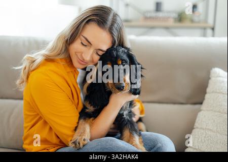 Glückliche junge kaukasische Frau mit langen blonden Haaren und lässigen Kleidern, in einem gelben Hemd, sitzt auf einer Couch im gemütlichen Wohnzimmer und umarmt ihren schwarz-braunen Hund mit einem liebevollen Ausdruck Stockfoto