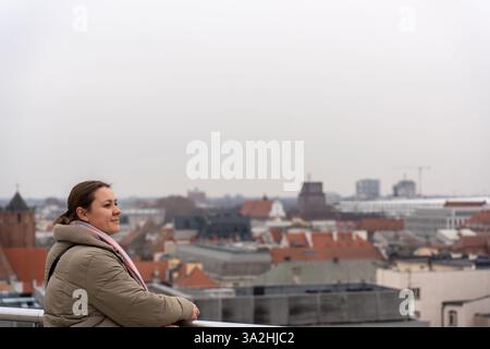 Lächelnde Frau in warmer Jacke und Schal, lehnt sich auf Geländer und genießt an einem bewölkten Tag einen Panoramablick auf die Stadt. Stockfoto