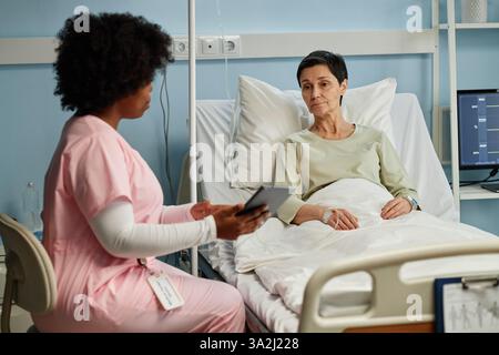 Schwester sitzt neben einem Bett, hält einen Notizblock und interagiert mit einem älteren Patienten, der im Bett eines Krankenhauszimmers liegt Stockfoto