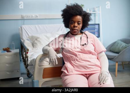 Porträt einer afroamerikanischen Krankenschwester, die auf dem Krankenhausbett sitzt und rosa Peelings mit Stethoskop um den Hals trägt. Der Raum verfügt über medizinische Geräte und ein Patientenbett im Hintergrund Stockfoto