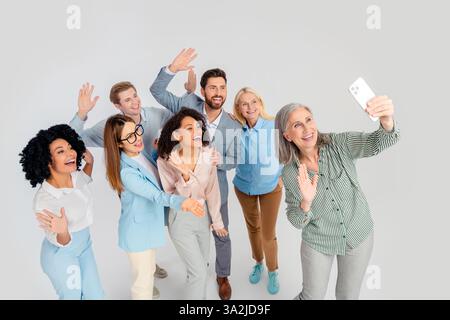 Verschiedene Geschäftsleute lächeln und winken, während sie zusammen mit grauem Hintergrund ein Selfie machen Stockfoto
