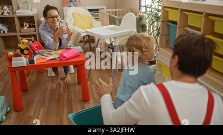 Mutter fotografiert Vater und Sohn, die zusammen in einem gemütlichen Wohnzimmer mit farbenfrohen Spielzeugen und warmer Beleuchtung spielen, um einen Moment der Familie festzuhalten Stockfoto