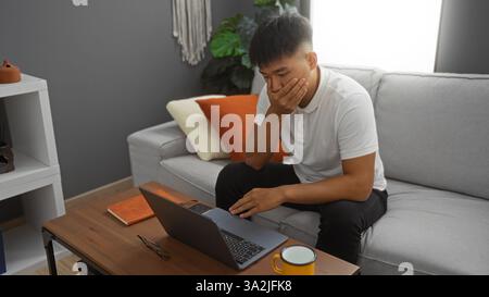 Der junge Mann scheint besorgt zu sein, während er in einem gemütlichen Wohnzimmer mit Hand an Mund auf einen Laptop blickt, was die Bedenken in Innenräumen mit moderner Einrichtung widerspiegelt. Stockfoto