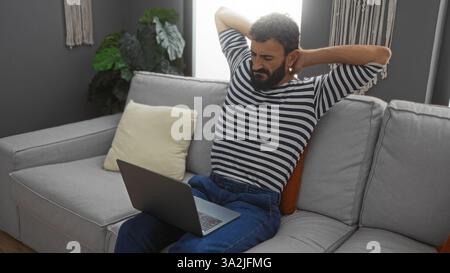 Junger hispanischer Mann, der sich auf einer Couch in einem Wohnzimmer mit einem Laptop entspannt und sich ausdehnt, während er auf den Bildschirm blickt, in einer komfortablen Apartmentumgebung. Stockfoto