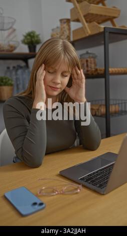 Frau, die im Büro sitzt und sich mit einem Laptop belastet fühlt, hält den Kopf mit Händen, die von Smartphone und Brille auf Holztisch umgeben sind. Stockfoto