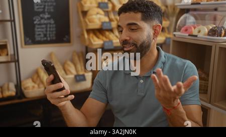Junger Mann, der in einer Bäckerei eine Videollamada macht, mit Brot und Gebäck im Hintergrund Stockfoto