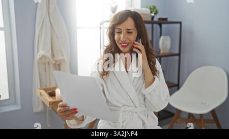 Schöne Frau mittleren Alters in einem weißen Bademantel, die am Telefon spricht und ein Dokument in einem gemütlichen, gut beleuchteten Badezimmer mit Stuhl und Muschel hält Stockfoto