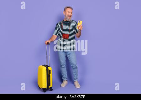 Ein stilvoller Seniorenmann mit Gepäck überprüft sein Smartphone vor einem leuchtenden lila Hintergrund, der auf Reisen reflektiert Stockfoto