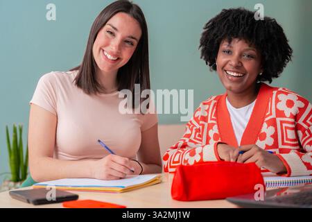 Zwei multikulturell lächelnde junge Studentinnen studieren zusammen an einem Schreibtisch und schauen in die Kamera Stockfoto