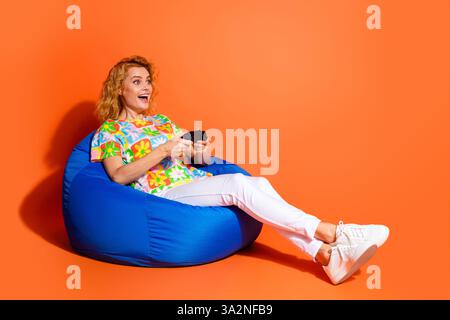 Fröhliche Frauen spielen in einem Sitzsack-Stuhl vor einem leuchtenden orangefarbenen Hintergrund und bringen Freude und Spannung in lässiger Sommermode zum Ausdruck Stockfoto