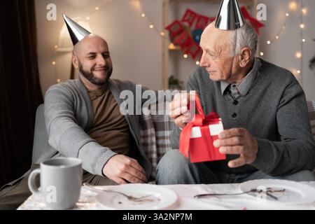Glückliche Familie Sohn mittleren Alters und älterer Vater in Partyhüten feiern Geburtstag zu Hause, Mann schenkt Dad Geschenkbox am Jahrestag Stockfoto