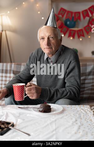 Porträt eines pensionierten Mannes mit Partyhut, der allein Geburtstag feiert und mit Traurigkeit in die Kamera schaut Stockfoto