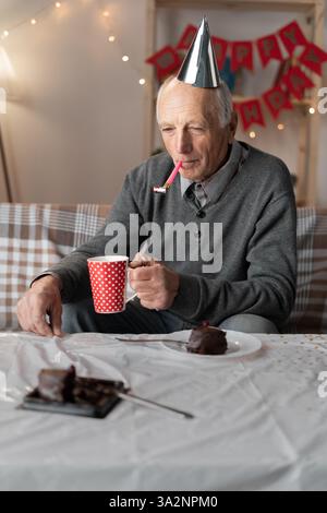 Unglücklicher pensionierter Mann mit Kegelhut mit Horn im Mund, der mit gelangweiltem Ausdruck wegschaut und seinen traurigen Geburtstag feiert. Stockfoto