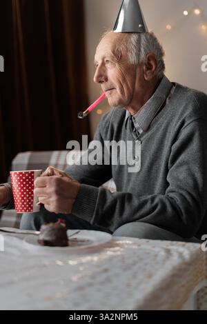 Unglücklicher pensionierter Mann mit Kegelhut mit Horn im Mund, der mit gelangweiltem Ausdruck wegschaut und seinen traurigen Geburtstag feiert Stockfoto