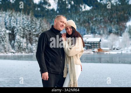 Paare stehen zusammen auf einem gefrorenen See in einer ruhigen Winterlandschaft mit schneebedeckten Bergen und immergrünen Wäldern, die entspannt und zufrieden ausstrahlen Stockfoto