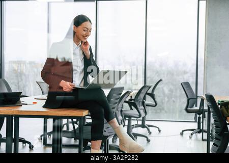 Auf dem Tisch sitzen, mit Laptop auf den Knien. Geschäftsfrau ist drinnen im Büro. Stockfoto
