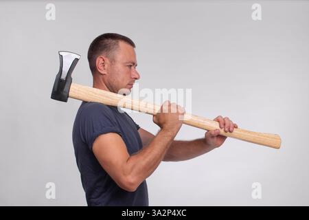 Ein Mann mit einer Axt steht vor einem grauen Hintergrund, in einer Seitenansicht erfasst. Das Bild betont die Stärke des Mannes und die Schärfe der Axt, wodurch eine kraftvolle und dynamische Komposition entsteht. Konzept Stockfoto