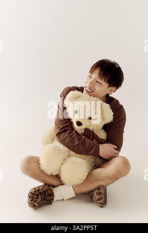 Ein hübscher junger asiatischer Mann lächelt hell, während er einen großen Teddybären in einer gemütlichen Umgebung umarmt. Stockfoto
