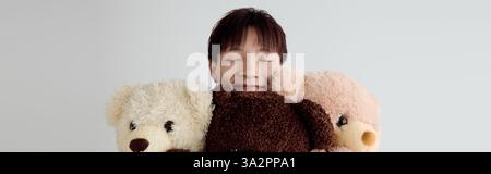 Ein junger, gutaussehender asiatischer Mann lächelt hell, während er drei entzückende Teddybären in einem gemütlichen Zimmer umarmt. Stockfoto