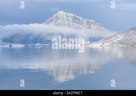Schnee bedeckt die Berge und Bäume, während der stille See die ruhige Winterlandschaft während des frühen Morgenlichts reflektiert. Stockfoto