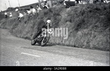 1955, Historic, ein Radfahrer auf der Hügelstrecke des Isle of man TT-Rennens, in Leder, mit einem traditionellen Helm und einer Gesichtsbrille, versammelten sich die Zuschauer auf der grasbewachsenen Piste und beobachteten das Rennen. Die Motorradrennen TT oder Tourist Trophy auf der Isle of man gehen auf das Jahr 1907 zurück. Stockfoto