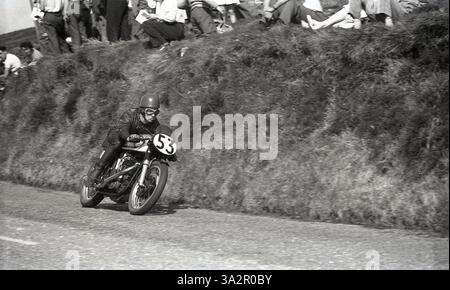 1955, Historic, ein Radfahrer auf der Hügelstrecke des Isle of man TT-Rennens, in Leder, mit einem traditionellen Helm und einer Gesichtsbrille, versammelten sich die Zuschauer auf der grasbewachsenen Piste und beobachteten das Rennen. Die Motorradrennen TT oder Tourist Trophy auf der Isle of man gehen auf das Jahr 1907 zurück. Stockfoto
