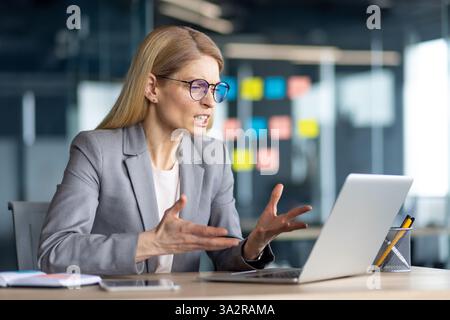 Eine Geschäftsfrau sitzt an ihrem Schreibtisch und führt online mit ihrem Laptop in einer modernen Büroumgebung eine hitzige Diskussion. Stockfoto