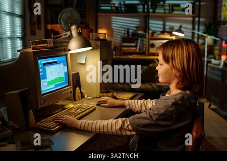 Kind sitzt am Schreibtisch mit einem alten Computer in einem gemütlichen, schwach beleuchteten Raum am Abend mit warmer Beleuchtung und verschiedenen Büchern und Dekorationen in den Regalen Stockfoto