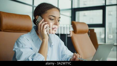 Eine junge Frau, die in einem modernen Büro multitaskt, mit einem Stift am Telefon telefoniert, fokussiert und mit einem Laptop an ihrem Arbeitsplatz engagiert wirkt Stockfoto