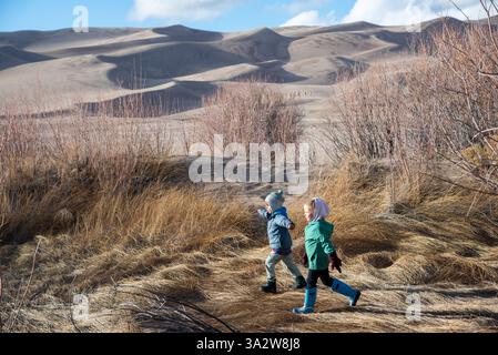 Zwei Kinder spielen im Gras mit den Großen Sanddünen im Hintergrund. Great Sand Dunes National Park, Colorado. Stockfoto