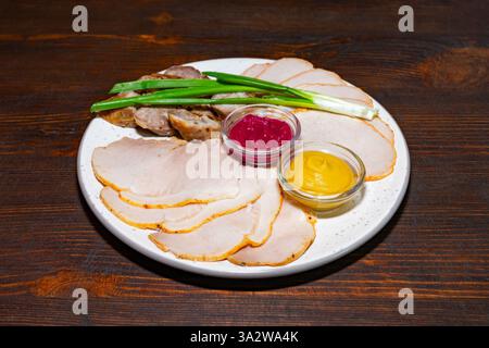 Eine Platte besteht aus dünn geschnittenem Fleisch, begleitet von Senf und einer süßen Sauce, garniert mit grünen Zwiebeln auf einer rustikalen Holzoberfläche. Stockfoto