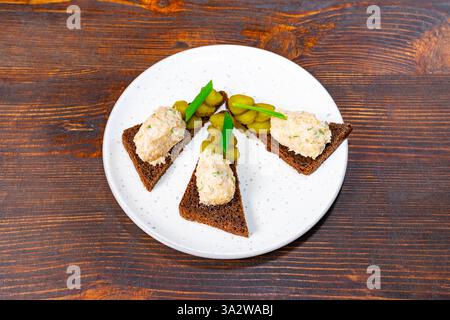 Kleine dreieckige Roggenbrotstücke, garniert mit cremigem Aufstrich und garniert mit Gurken sind wunderschön auf einem weißen Teller angeordnet, was einen so hervorhebt Stockfoto