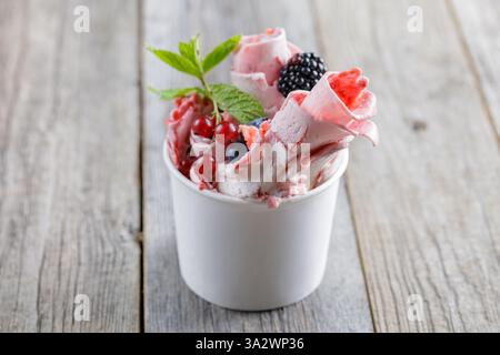 Köstliches, gerolltes Eis in einer Tasse, garniert mit frischen Beeren, einem Schokoladen-Waffelstab und Obstgarnituren, serviert auf einem rustikalen Holztisch Stockfoto