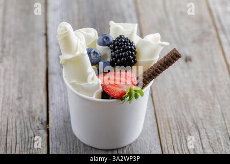 Köstliches, gerolltes Eis in einer Tasse, garniert mit frischen Beeren, einem Schokoladen-Waffelstab und Obstgarnituren, serviert auf einem rustikalen Holztisch Stockfoto