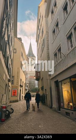 Ein Paar, das mit einem Hund in einer engen Gasse läuft. Die Gasse ist von Gebäuden gesäumt und hat einen gemauerten Gang Stockfoto