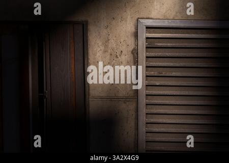 Eine abstrakte Komposition aus einer Lüftungsöffnung und einer halb verborgenen Tür auf einer rohen Betonwand, umgeben von tiefen Schatten, einer geheimnisvollen und stimmungsvollen Atmosphäre Stockfoto
