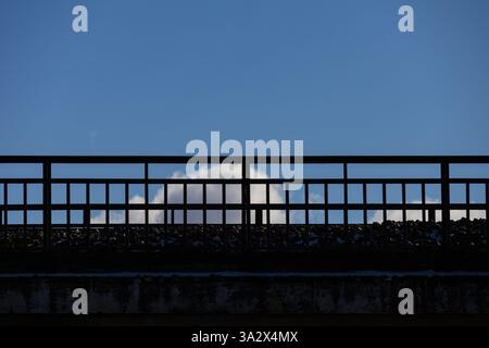 Von unten fotografiert, bildet eine Zementbrücke mit Metallgeländern sich kreuzende Quadrate und Rechtecke in Silhouette, die vor einem klaren blauen Himmel stehen Stockfoto