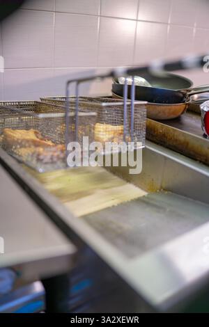 Mit Samosas gefüllte Körbe aus Metall, die Öl in eine Fritteuse in einer gewerblichen Küche geben, was die Zubereitung eines traditionellen indischen Desserts unterstreicht Stockfoto