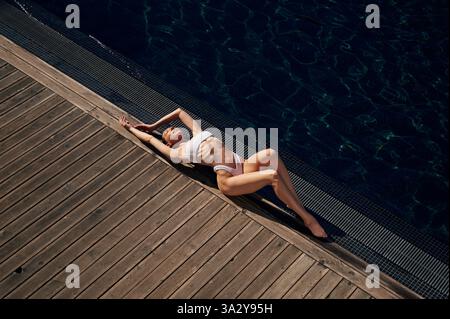 Sich hinlegen und das Sonnenlicht genießen. Frau in weißem Bikini ist in der Nähe des Schwimmbades. Stockfoto