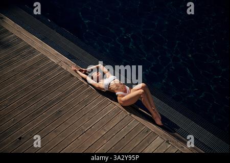 Sich hinlegen und das Sonnenlicht genießen. Frau in weißem Bikini ist in der Nähe des Schwimmbades. Stockfoto